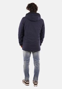 Giacca puffer blu navy con cappuccio, caratterizzata da una texture liscia, polsini elasticizzati e lunghezza fino a metà coscia. Indossata sopra jeans azzurri chiari e sneakers marroni.