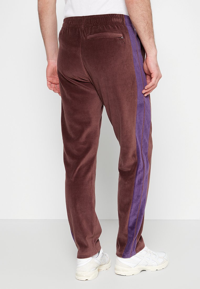Jogging en velours brun avec des rayures latérales violettes, taille élastique et poche zippée à l'arrière. Texture lisse, design à jambe fuselée.