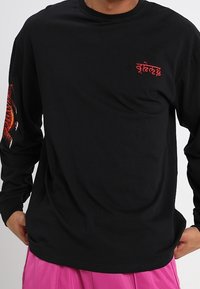 Mustapaita, jossa on pitkät hihat, valmistettu puuvillasta. Vasemmassa hihassa on punainen kuvio ja rintakehässä brodeeraus. Sujuva pinta.