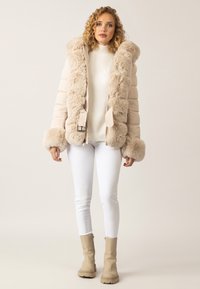 Beige gepolsterter Mantel mit einem Kragen und Manschetten aus Kunstpelz, frontalem Reißverschluss und Gürtelakzent. Kombiniert mit weißen skinny Jeans und beige Stiefeletten.
