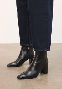 Botas negras de cuero hasta el tobillo con tacones cuadrados, combinadas con jeans de pierna recta azul oscuro, de pie sobre un suelo de concreto liso.