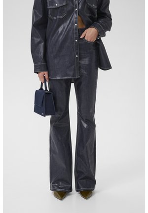 Personne portant une chemise en denim sombre et brillante avec un pantalon évasé, tenant un petit sac à main bleu marine, une main dans la poche, debout sur fond blanc.