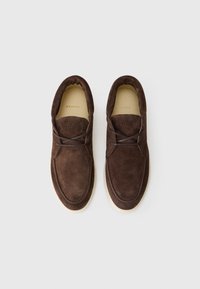 Pantofi slip-on din suede maro, cu o formă rotundă, având o talpă de profil redus și cusături subtile. Interiorul este căptușit într-o culoare deschisă.
