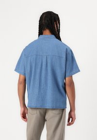 Short-sleeve denim shirt in light blue with a back yoke, pleat detail, and a collar. Material appears soft with a smooth texture.