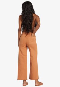 Donna con lunghi capelli mossi è di spalle, indossa pantaloni larghi arancioni, sandali e un top senza schiena con una mano in tasca sul retro.