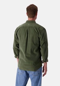 Camicia verde a maniche lunghe con colletto e maniche arrotolate, dotata di un sottile dettaglio del logo sul retro. Indossata con jeans blu.