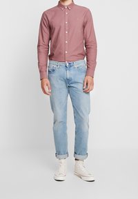 Långärmad, ljusrosa skjorta med knappar i vitt, i kombination med ljusblå uppvikta jeans och vita högskurna sneakers.