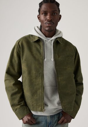 Homme aux cheveux tressés portant une veste texturée verte par-dessus un sweat à capuche gris avec cordons, debout devant un fond clair uni.