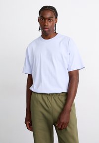 T-shirt de algodão azul claro com decote redondo e mangas curtas, apresentando um logótipo em relevo. Combinado com calças olivais com cordão.