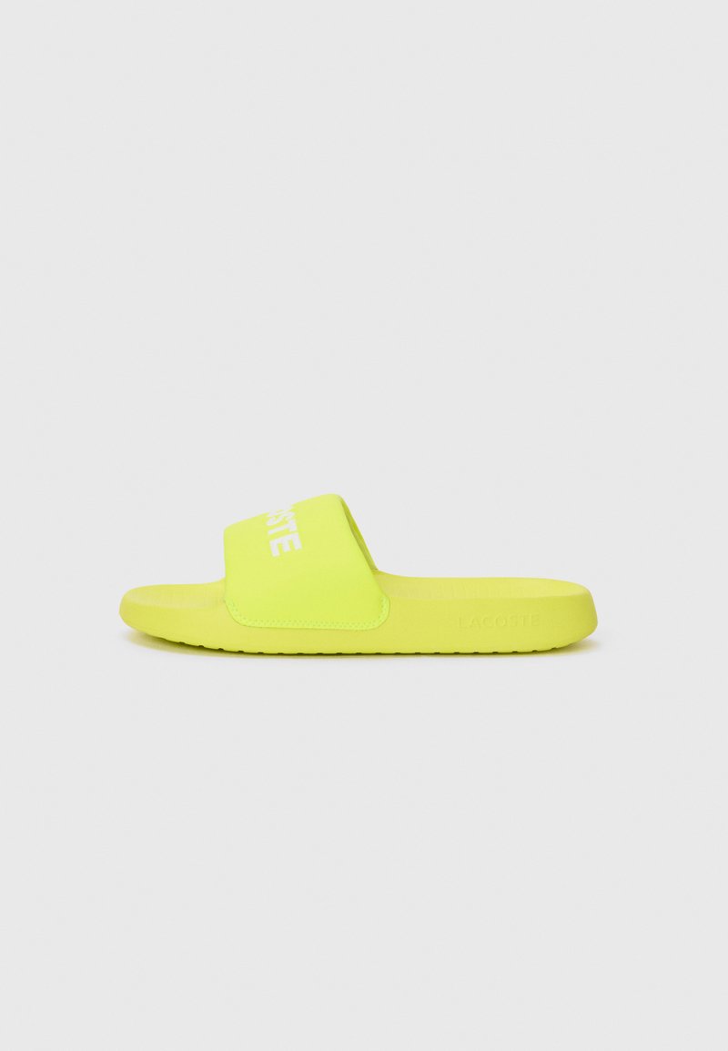 Spilgti dzelteni Lacoste slaidu sandales ar plašu siksnu un iegravētu logotipu, skatīts no sāniem uz vienkārša balta fona.