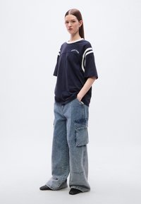 Μπλε ναυτικό φαρδύ t-shirt με λευκές ρίγες, βαμβακερό ύφασμα και κεντημένο λογότυπο, συνδυασμένο με ανοιχτό μπλε παντελόνι cargo με φαρδιά μπατζάκια και τσέπες.