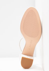 Chaussure à talons hauts blanche avec une tige en cuir lisse, une sangle de cheville transparente et une semelle en caoutchouc marron clair présentant un motif de grip texturé.