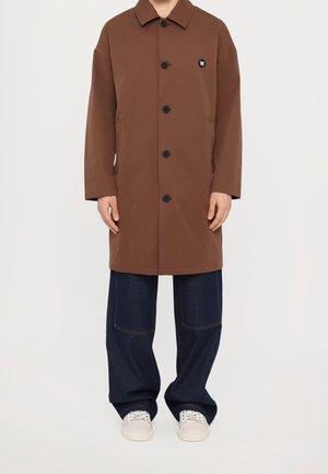 Mężczyzna ubrany w brązowy płaszcz na guziki, ciemne luźne dżinsy i białe trampki, stojący na tle jednolitego jasnego tła.