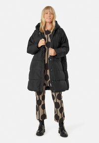 Cappotto imbottito nero con cappuccio, caratterizzato da un design trapuntato. Indossato sopra un outfit beige a motivi. Stivali neri con lacci completano il look.