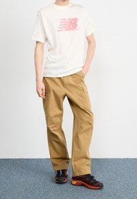 Camiseta corta de manga con logo gráfico rojo, combinada con pantalones cargo color marrón con bolsillos laterales y zapatillas deportivas negras y rojas.