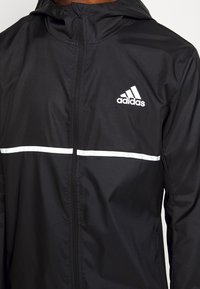 Svart Adidas zip-up jacka med huva, vit rand över bröstet och Adidas-logotyp på vänster bröst.