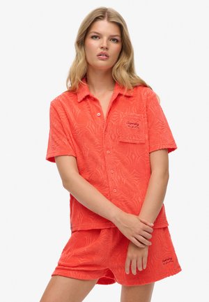 Camisa de manga corta color coral brillante y pantalones cortos a juego en tela texturizada con patrones sutiles de ondas; incluye bolsillo frontal y logo bordado.