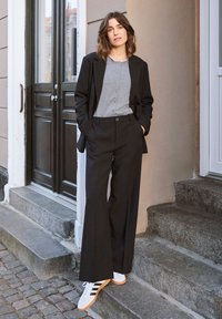 Czarny dopasowany żakiet i szerokie spodnie noszone na szarej koszuli z guzikami. Białe sneakersy z czarnymi paskami. W tle kamienne schody i drewniane drzwi.