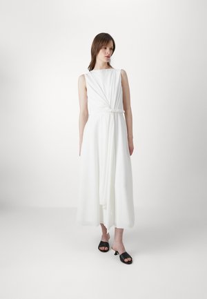 Witte, mouwloze jurk met een geknoopte taille, vloeiende stof en lange zoom. Zwarte open teen hakken maken de look compleet.