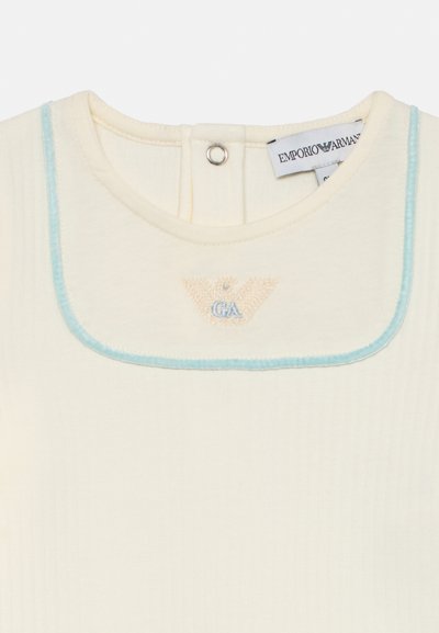 Kremfarget ribbet stofftopp med lyseblå kragekant, brodert logo i beige og blått, og med trykknapplukking bak.