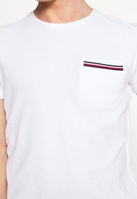 Biała bawełniana koszulka z okrągłym dekoltem i krótkimi rękawami, z kieszonką na piersi ozdobioną czerwonymi i czarnymi paskami.