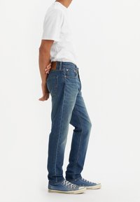 En person klädd i blå jeans, vit t-shirt och blå sneakers står med händerna knäppta bakom ryggen mot en enkel vit bakgrund.