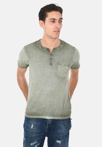 Maglietta henley a maniche corte verde chiaro con finitura testurizzata, placca a tre bottoni e tasca sul petto. Indossata con jeans blu strappati.