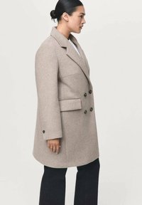 Manteau beige en mélange de laine à double boutonnage avec un col cranté, de forme rectangulaire, poches latérales et boutons foncés, porté sur un pantalon bleu marine.