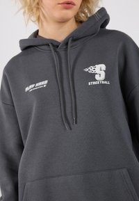 Mørkegrå hettegenser med frontlomme, med hvit tekst og grafikk: "PLAY HARD" og "STREETBALL" med flammedetaljer.