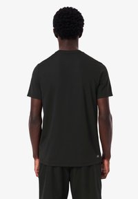 Man med kort lockigt hår klädd i en enkel svart t-shirt med kort ärm och svarta byxor, stående vänd bort från kameran mot en vit bakgrund.