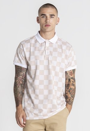 Ung mand med kort hår og tatoverede arme iført en hvid og beige ternet poloshirt og beige bukser, stående foran en ensfarvet baggrund.