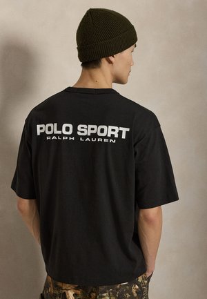 Mężczyzna w czarnej koszulce Polo Sport Ralph Lauren i oliwkowej dzianej czapce, pokazany tyłem na jednolitym beżowym tle.