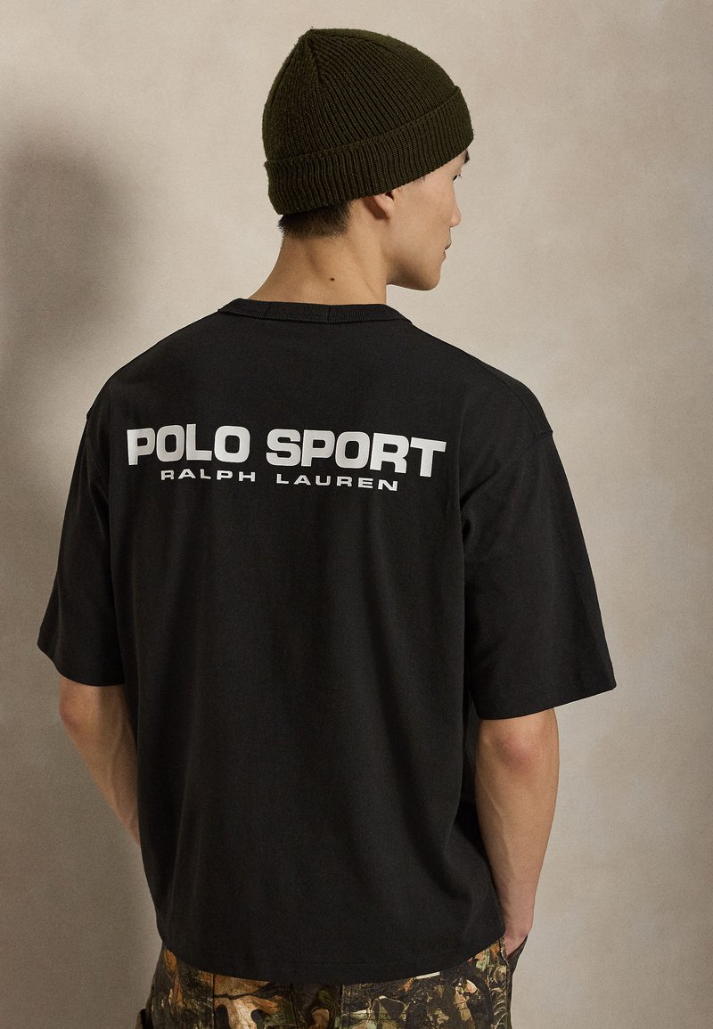 Άνδρας που φοράει μαύρο μπλουζάκι Polo Sport Ralph Lauren και ελιά πλεκτό σκουφάκι, με την πλάτη προς τα έξω σε μονόχρωμο μπεζ φόντο.