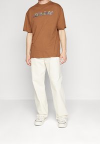 Brun bomullströja med blommönster över bröstet, kombinerad med vita raka byxor och vita sneakers.