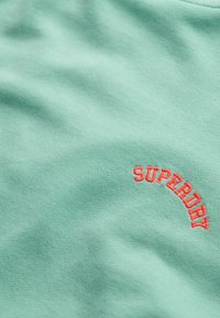 Lichtgroene katoen stof met een gladde textuur; heeft een rood geborduurd "SUPERDRY" logo in een boogvorm.
