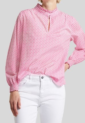 Femme portant un chemisier rose à manches longues avec une petite fente avant et un pantalon blanc, debout avec une main sur la hanche.