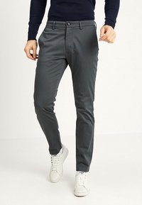 Grå, slim-fit byxor med en slät textur, dragkedja och knappstängning, kombinerade med vita sneakers och en mörk tröja.