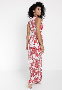 Lang, mouwloos jurkje van witte stof met rode paisley patronen, met een getailleerde bodice en een vloeiende rok, voorzien van een ronde halslijn.