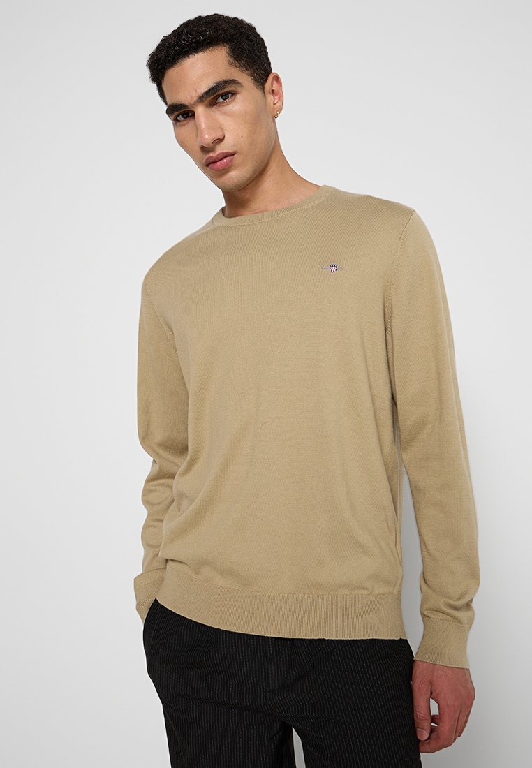 Beige Strickpullover mit Rundhalsausschnitt, ausgestattet mit einem kleinen, bunten Logo auf der linken Brust. Strukturierter Stoff und lockere Passform.