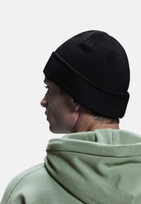 Un bonnet noir à côtes porté de travers sur la tête d'une personne, associé à un sweat à capuche vert clair. Le tissu semble épais et texturé.