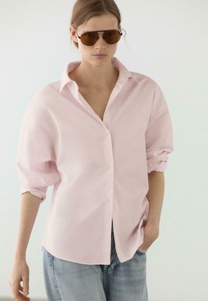 Kobieta nosząca oversize jasnoróżową koszulę zapinaną na guziki, podwinięte rękawy, jasnoniebieskie dżinsy i brązowe okulary przeciwsłoneczne, stojąca z jedną ręką w kieszeni.