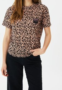 Tričko s leopardím vzorem v světle růžové barvě s černými skvrnami; kulatý výstřih, krátké rukávy a přední kapsa s detailní etiketou. Stylizováno s černými kalhotami.
