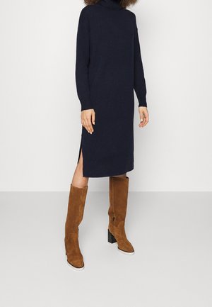 Frau trägt ein marineblaues, langärmliges Strickkleid mit Seitenschlitzen und kniehohe braune Wildleder-Stiefeletten mit Absatz vor einem schlichten Hintergrund.