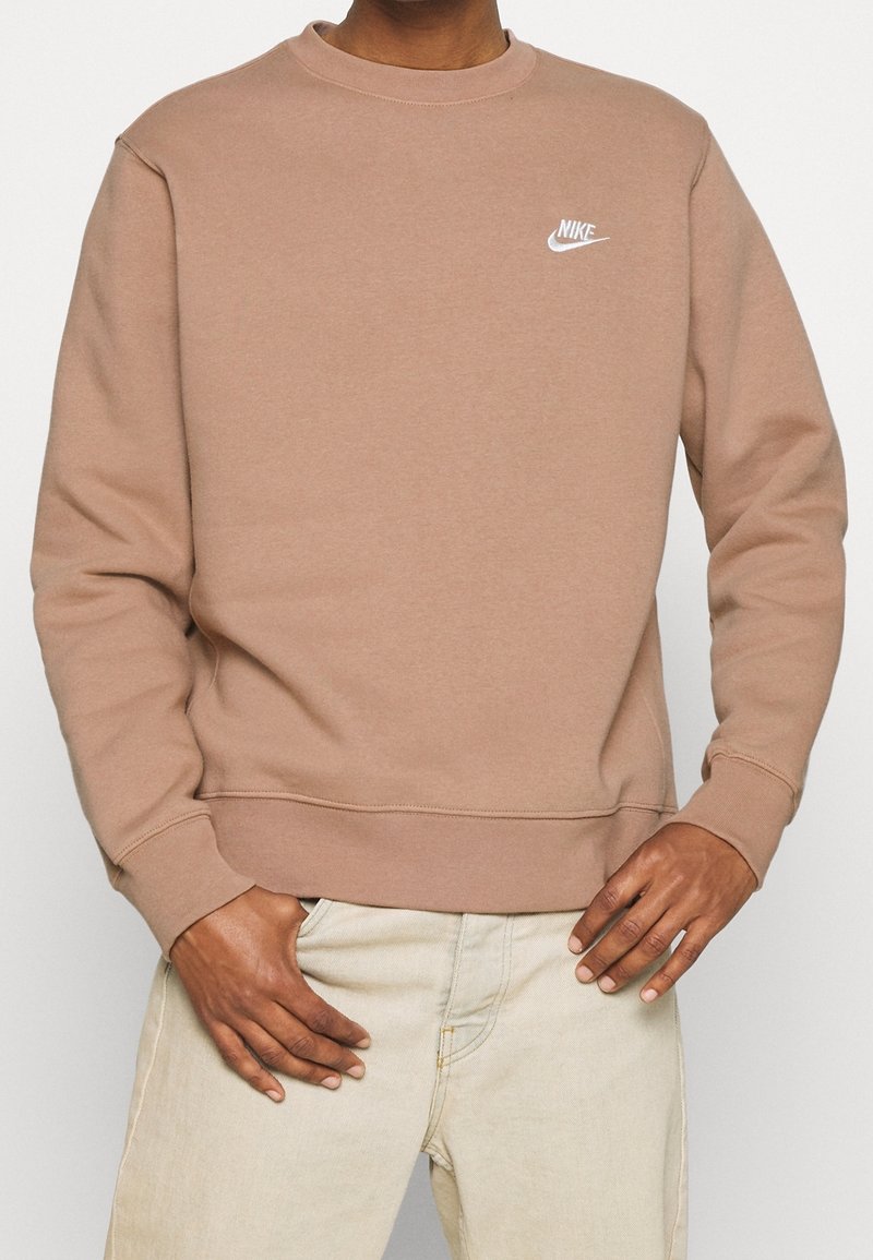 Personne portant un sweat Nike marron clair et un pantalon beige, debout, les mains posées près des poches, devant un fond uni.