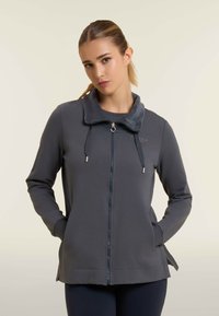 Giacca grigia con zip e colletto sicuro, realizzata in un tessuto morbido, dotata di cordoncini regolabili e tasche laterali. Dettaglio di branding discreto incluso.