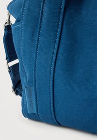 Γωνία από μπλε υφασμάτινη τσάντα με ρυθμιζόμενο λουράκι και ετικέτα Calvin Klein ορατή πάνω σε ομοιόμορφο φόντο.