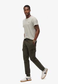 Grijze katoenen t-shirt, olijfgroene cargobroek met zijzakken, en witte sneakers met zwarte accenten. Gouden horloge om de pols, staande pose.