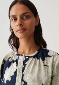 Jonge vrouw met lang donker haar die een marineblauwe en beige blouse met bloemenpatroon draagt, met een ronde halslijn en knopen.