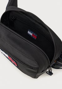 Sort stof skuldertaske med lynlåslommer, der har et farverigt logo-patch og et tekstureret design på stroppen.