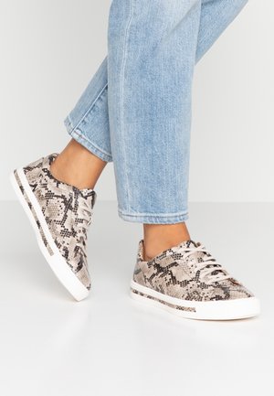 Person iført lyseblå jeans og lave sneakers med beige og sort slangehudsmønster og hvide såler.